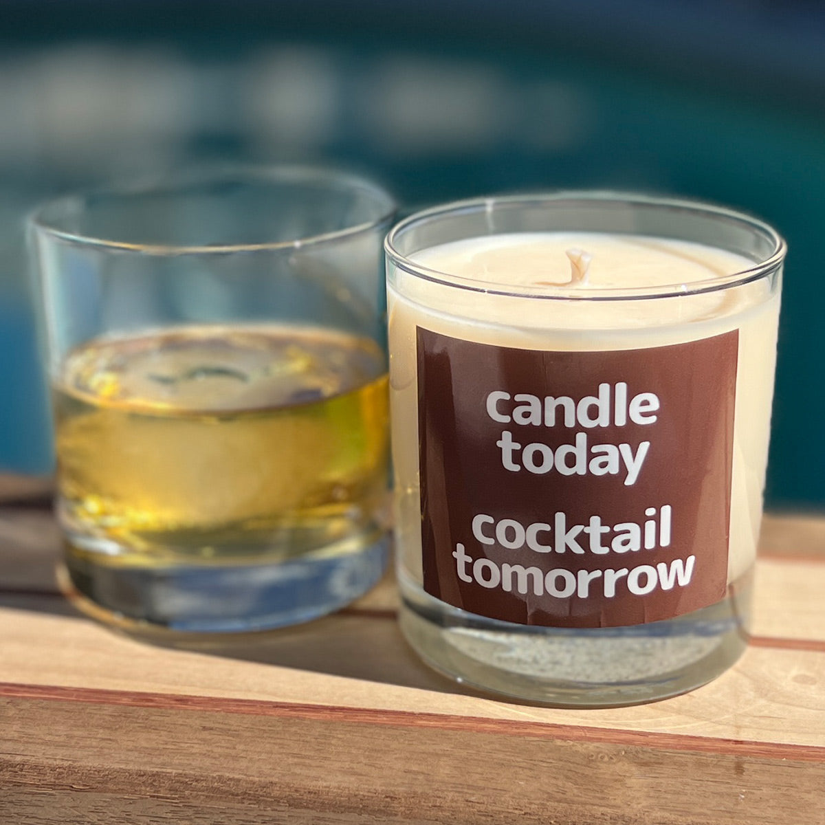Candle labeled 'candle today, cocktail tomorrow' next to a glass of bourbon on a wooden surface. Shows the dual purpose of these candles also being a rocks glass. 
