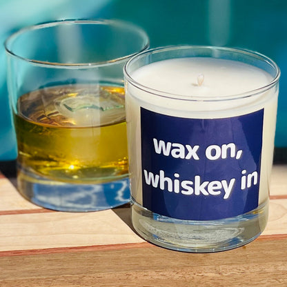 Candle labeled 'wax on, whiskey in' next to a glass of whiskey on a wooden surface.