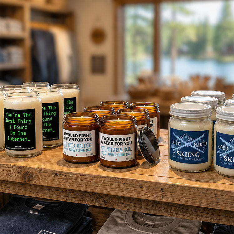 A display of scented candles in glass jars on a wooden shelf inside a retail store. The candles include white wax in amber and clear glass jars with humorous labels such as 'You're the Best Thing I Found on the Internet' and 'I WOULD FIGHT & BEAR FOR YOU'.