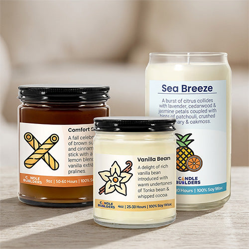 Three Candle Builders scented candles in glass jars of different sizes on a light wooden surface with a blurred indoor background. The candles include Comfort Candles, Vanilla Bean, and Sea Breeze, with black lids and white labels showing illustrations of spices and flowers.