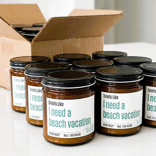 Several amber glass jars with black lids and white labels reading 'Smells Like I need a beach vacation' are arranged on a white surface, with an open cardboard box in the background.